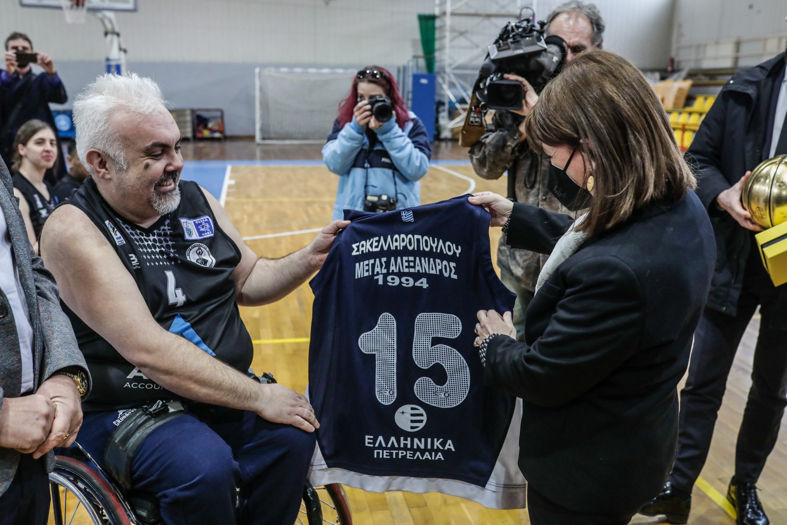 Η Πρόεδρος της Δημοκρατίας Κατερίνα Σακελλαροπούλου δέχεται δώρο μία φανέλα με το όνομά της μετά το τέλος του φιλικού αγώνα μπάσκετ ενηλίκων με αμαξίδια, που παρακολούθησε μεταξύ των ομάδων «Μέγας Αλέξανδρος» και «ΑΣΑΘ ΑΡΗΣ», στο κλειστό Δημοτικό Αθλητικό Κέντρο Πανοράματος, το Σάββατο 19 Μαρτίου 2022. 
