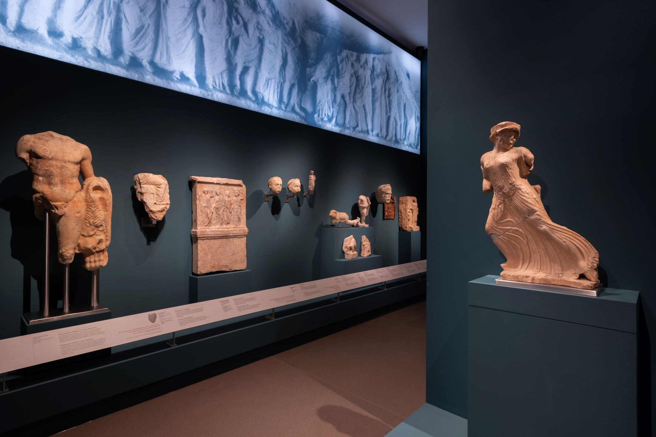Η «Φεύγουσα» Κόρη, εμβληματικό έκθεμα του Αρχαιολογικού Μουσείου Ελευσίνας και στην ίδια αίθουσα η επιγραφή για την γεφύρωση της λίμνης των Ρειτών (σήμερα λίμνη Κουμουνδούρου